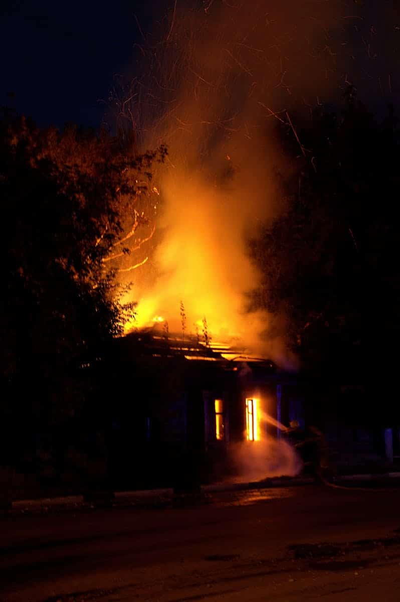Firefighters battle a raging house fire at night, with smoke and flames spiraling upward.