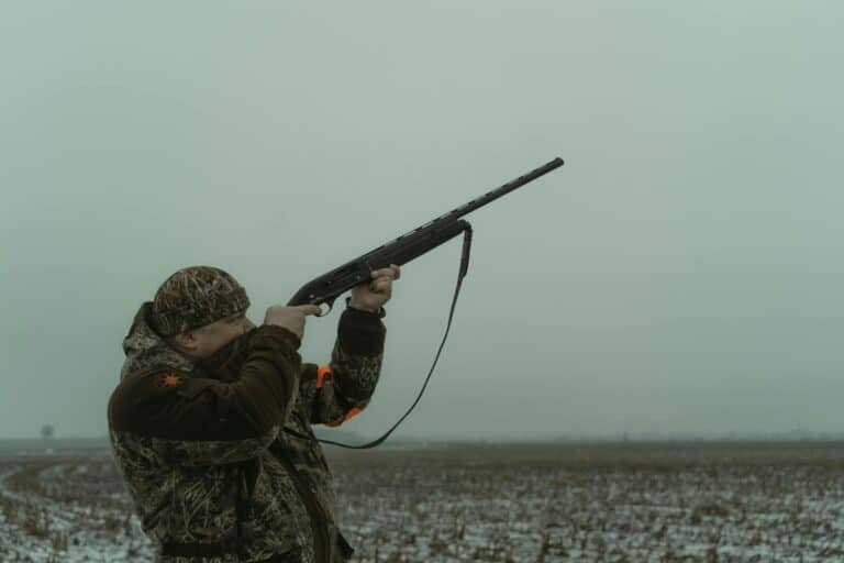 A hunter in camouflage aims a rifle in a foggy open field, showcasing outdoor hunting.