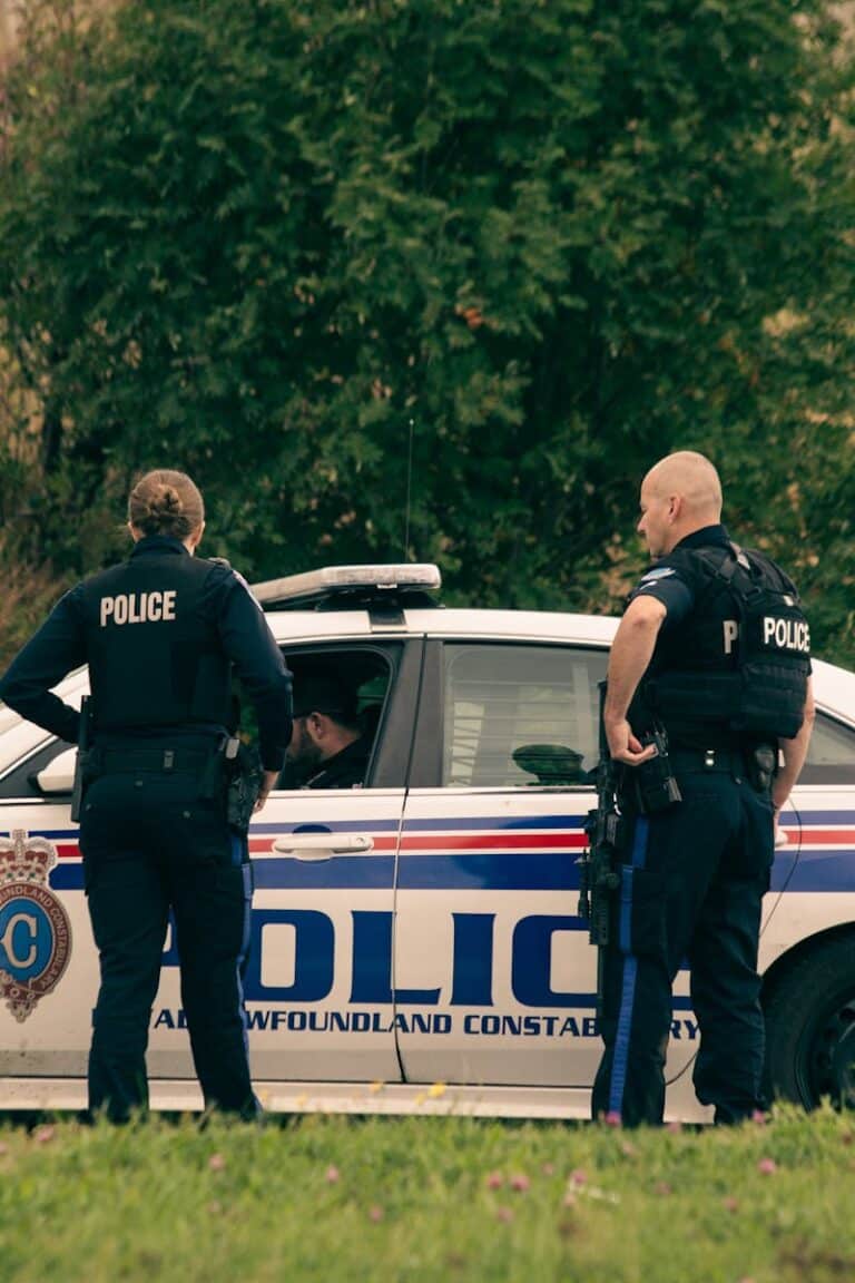 Back view of unrecognizable policewoman with male partner in uniform standing on meadow against automobile and tree