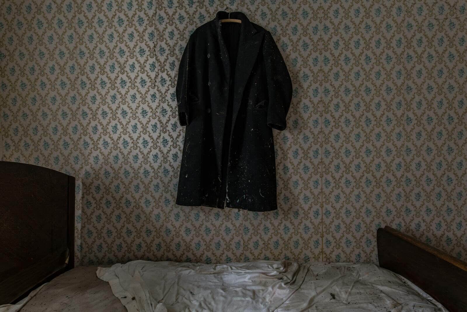A vintage black coat hangs on a wallpapered wall above an unkempt bed in an old-fashioned bedroom.
