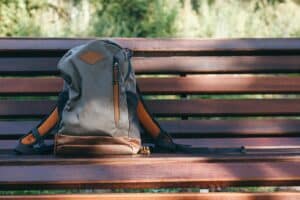 A brown and gray backpack resting on a wooden park bench in a sunny outdoor setting.