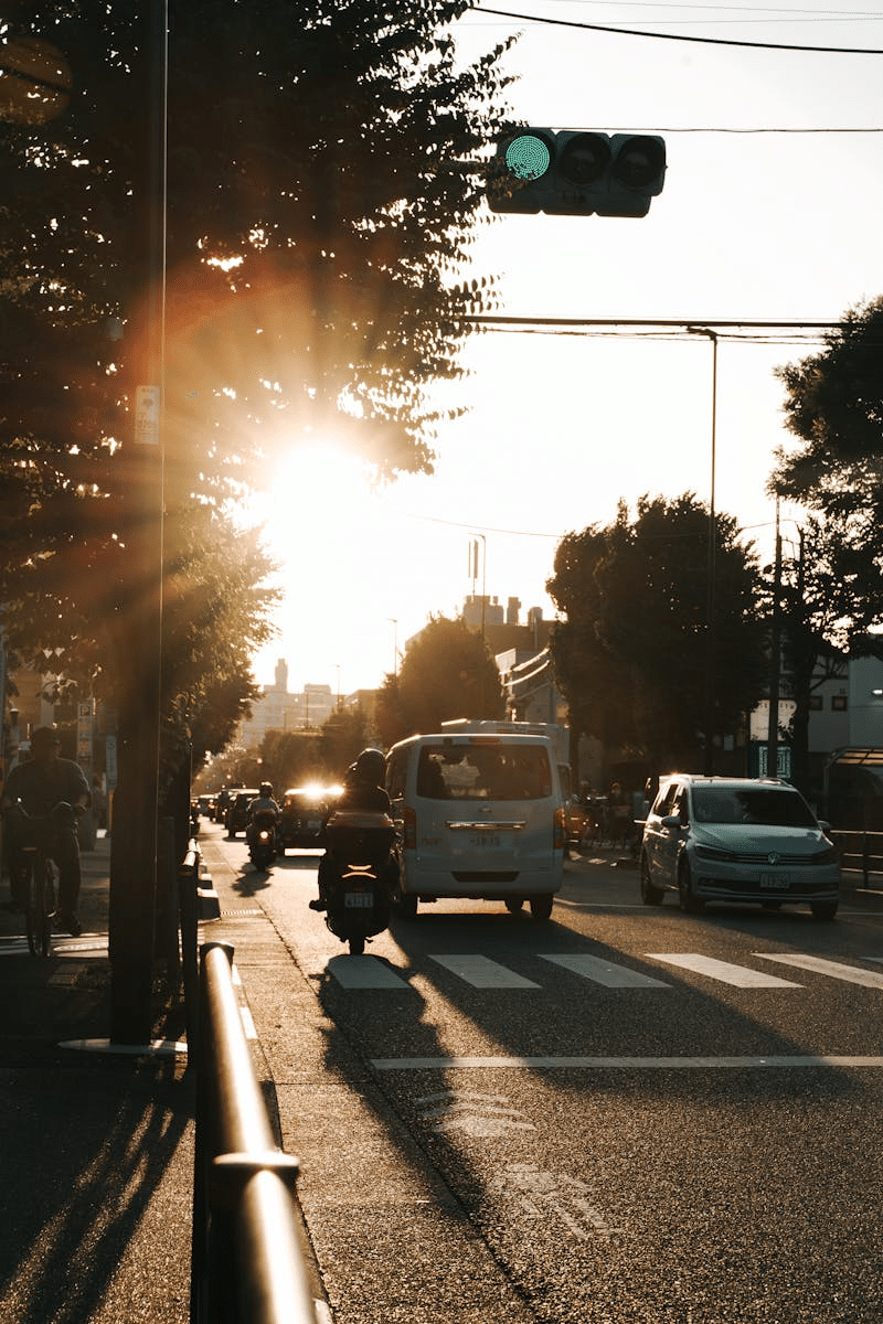 Cars and motorcycles drive on a city road, silhouetted by the rising sun.