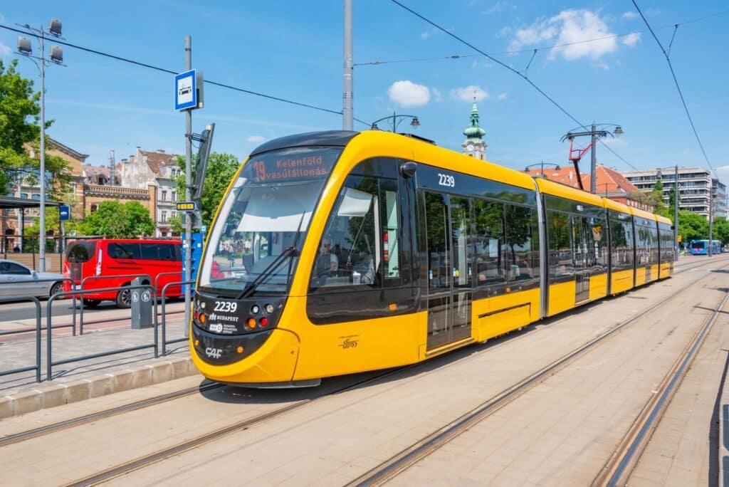 A yellow tram passes through a bustling city street on a bright day. Features urban architecture and transit.