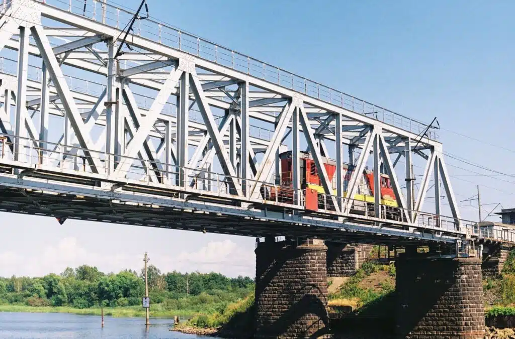 A red train crosses a steel bridge over a tranquil river on a clear day.